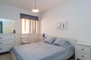 A bedroom with a bed, a nightstand, a lamp, a chair, a dresser, a window with blinds, and a wall-mounted light fixture.