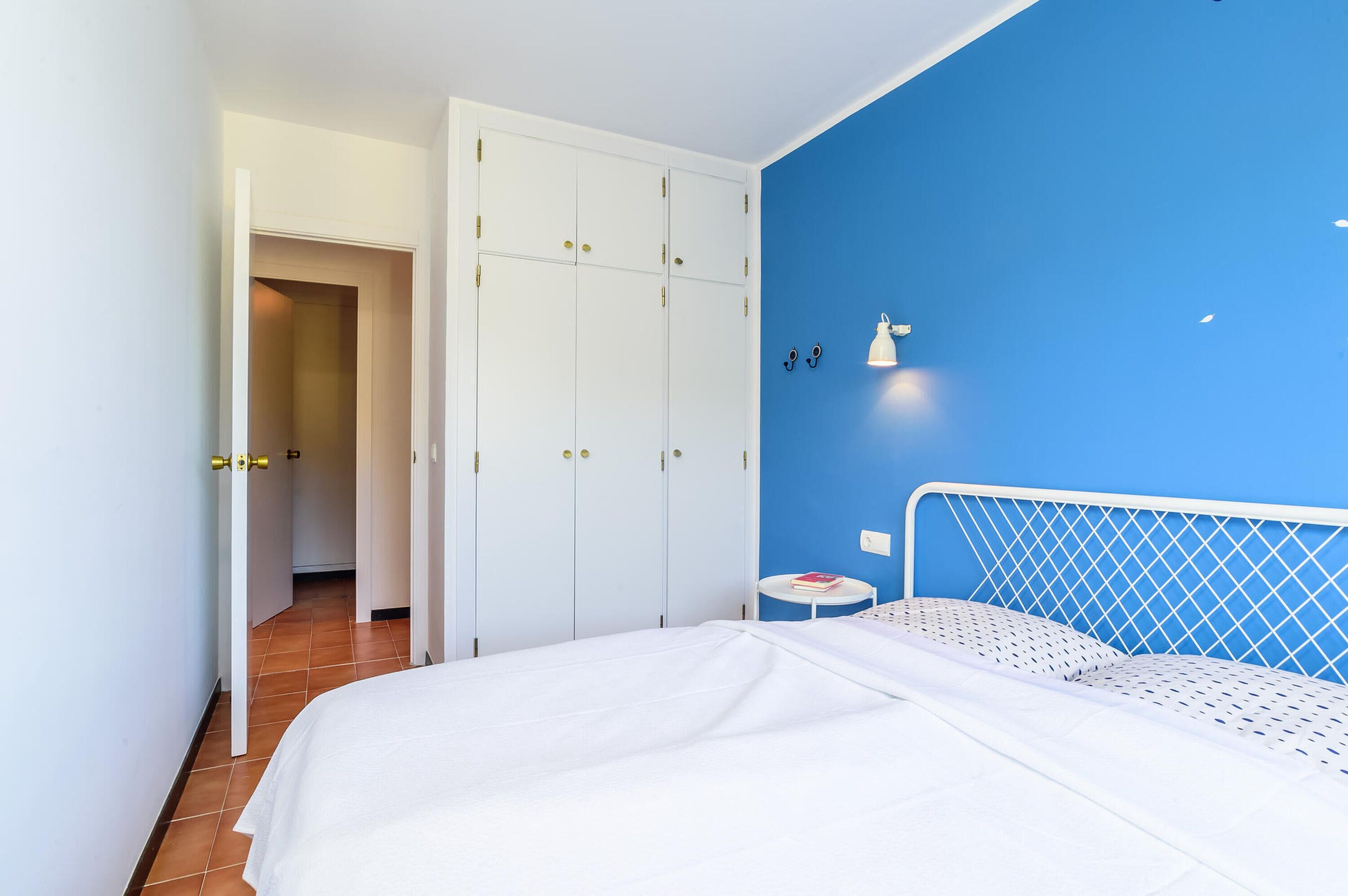 A bedroom with a bed, a wardrobe, a small table, and a wall-mounted lamp.