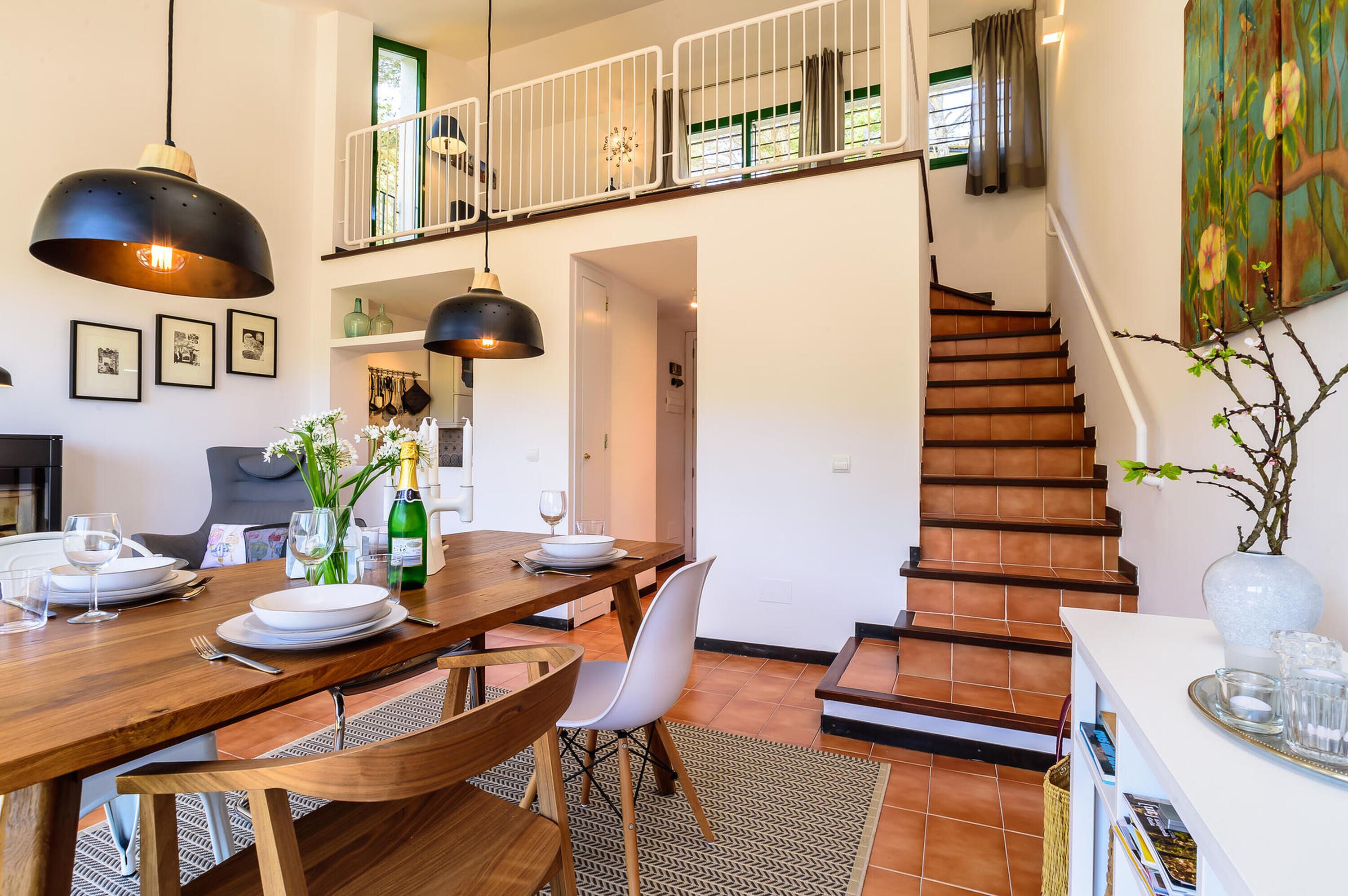 A dining area with a wooden table set for four, a staircase, and a painting on the wall.