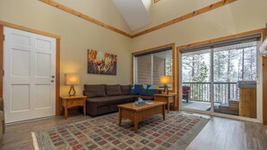 A cozy living room with a sofa, coffee table, and lamps. There is a large window with a view of trees and a balcony.
