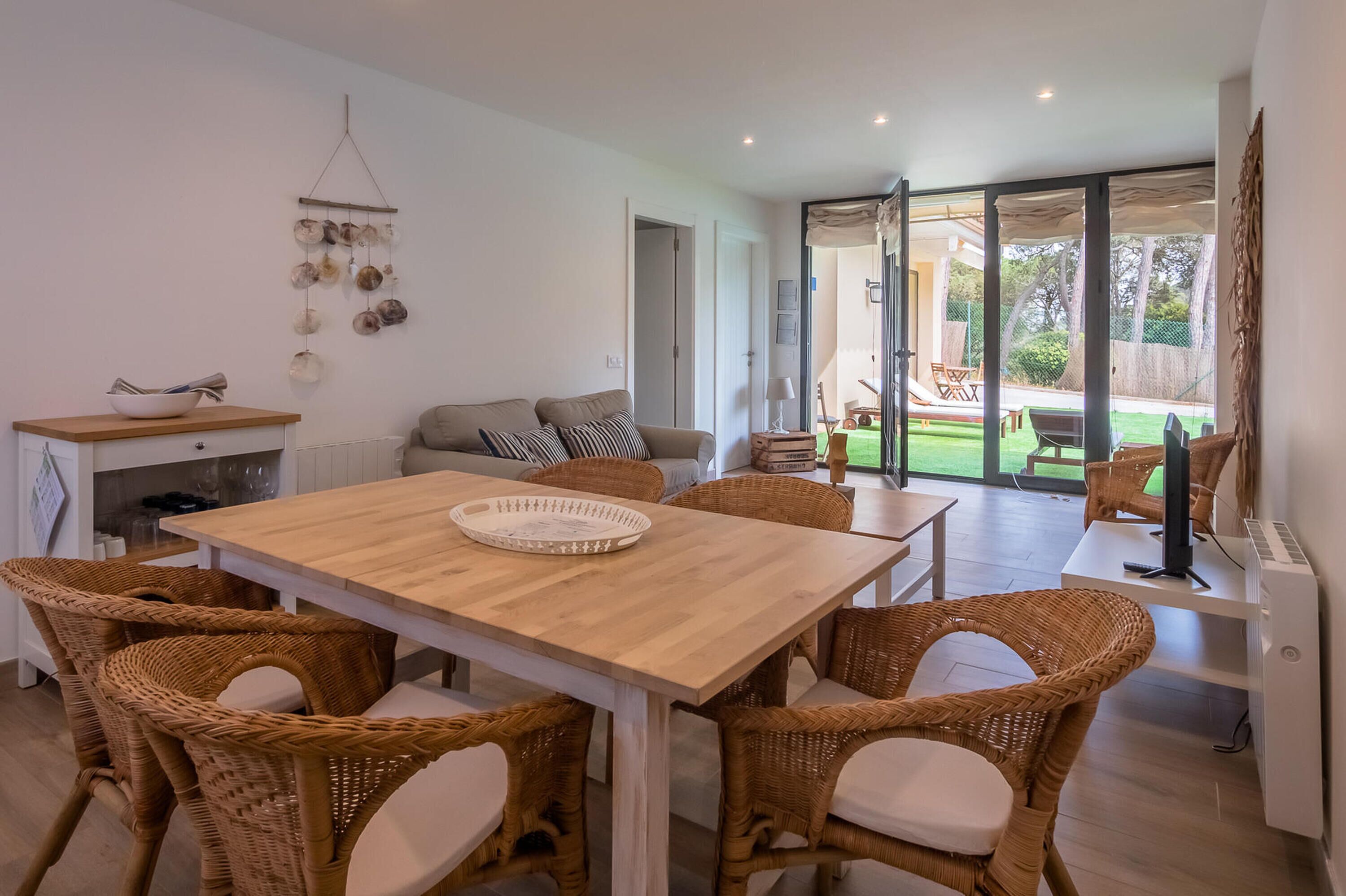 Una sala de estar moderna con una mesa de comedor de madera, sillas de mimbre, un sofá y un gran ventanal corredero que da a un espacio exterior.