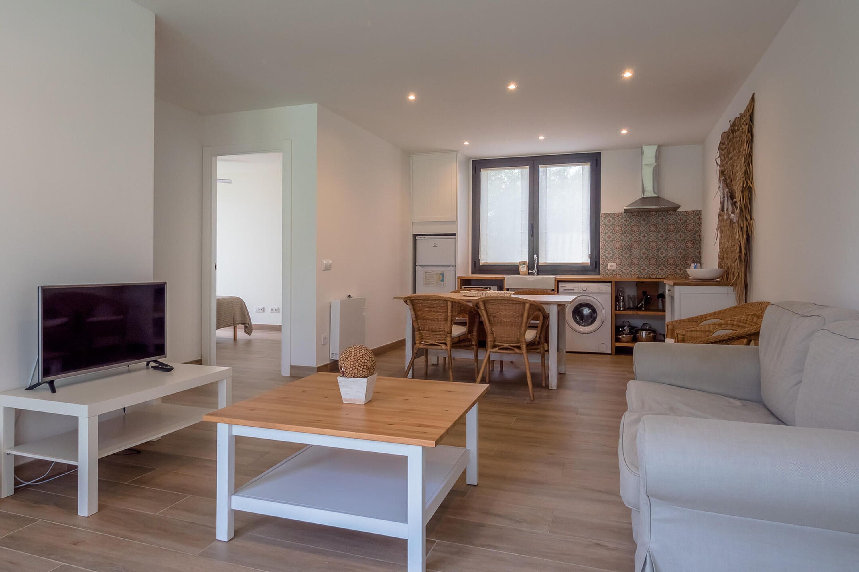 Un soggiorno moderno con televisione, un tavolino in legno e un divano. C'è un'area pranzo con sedie e un tavolo, e una cucina con una finestra e una lavatrice.