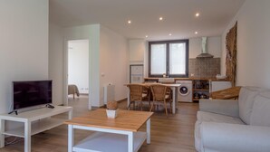 A modern living room with a TV, a wooden coffee table, and a sofa. There is a dining area with chairs and a table, and a kitchen with a window and a washing machine.
