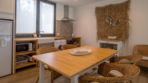 A compact kitchen with a wooden table and wicker chairs, a washing machine, a microwave, and a large woven tapestry on the wall.