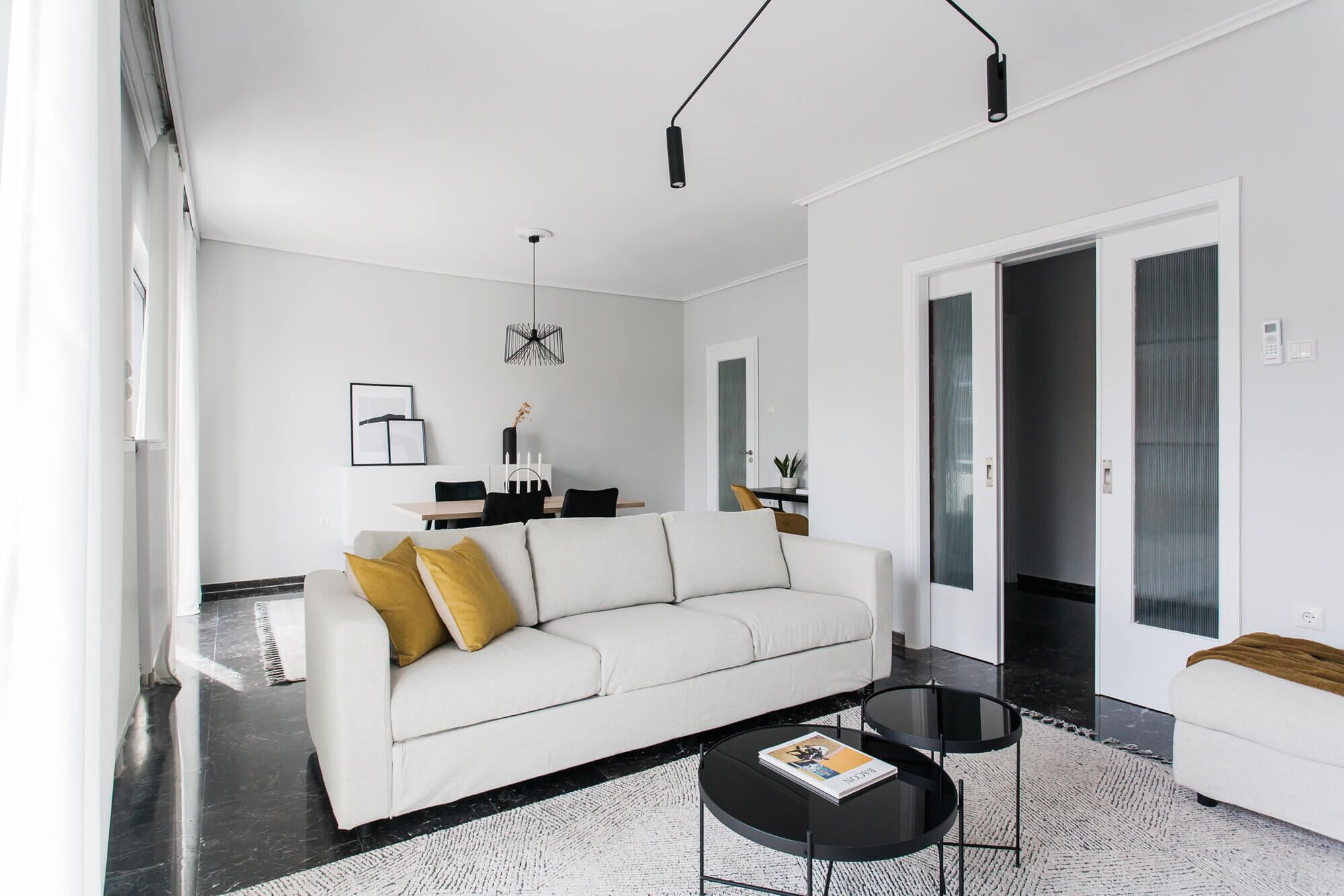 A modern living room with a white sofa, two black coffee tables, and a dining area with a white table and chairs.