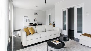 Ein modernes Wohnzimmer mit einem weißen Sofa, zwei schwarzen Couchtischen und einem Essbereich mit Tisch und Stühlen in Weiß.
