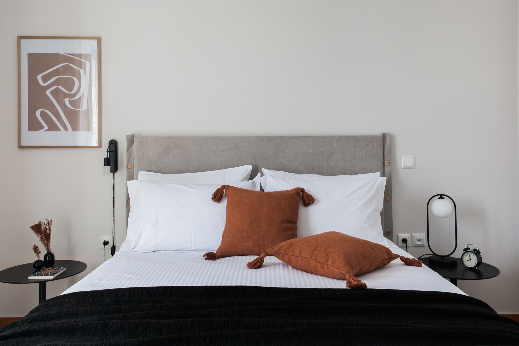 Un lit bien fait, avec une tête de lit grise, des oreillers blancs et bruns, et un plaid noir. On y trouve une petite table ronde avec un livre et des objets décoratifs, une lampe fixée au mur et, au-dessus du lit, une œuvre d'art abstraite encadrée.