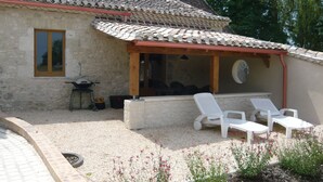A stone building with a tiled roof, a covered patio with white lounge chairs, and a grill outside. - Luxury gite for adult couples, peaceful, open views, large swimming pool (Boisse)