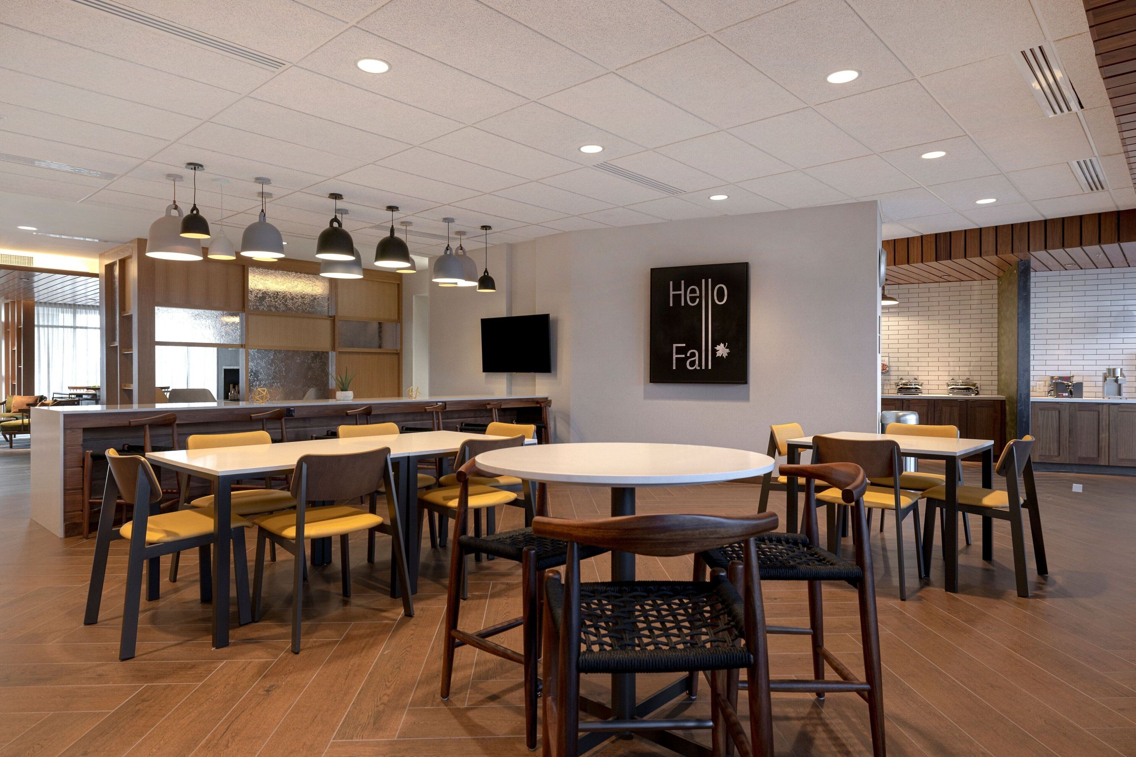 A modern dining area with multiple tables and chairs, a kitchen in the background, and a 'Hello Fall' sign on the wall.