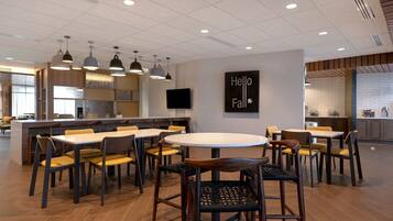 A modern dining area with multiple tables and chairs, a kitchen in the background, and a 'Hello Fall' sign on the wall.