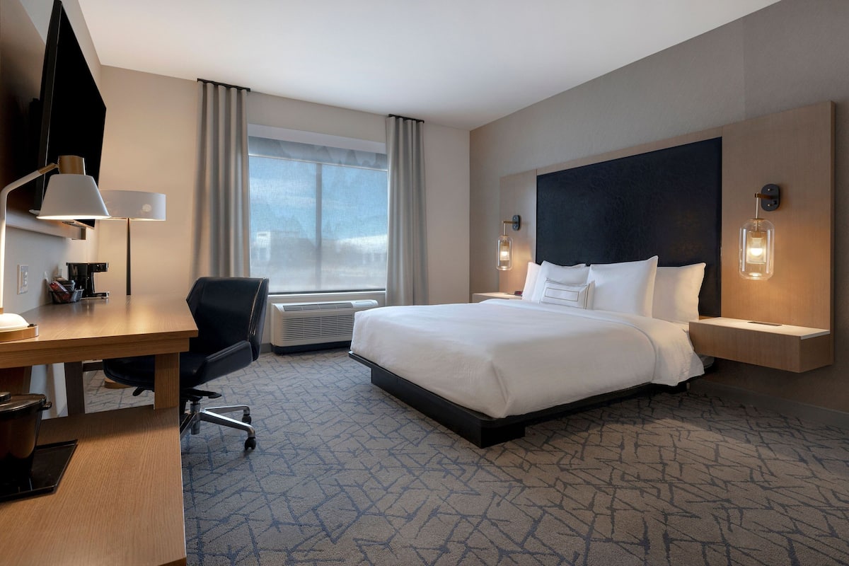 Una habitación de hotel con una cama grande, un escritorio con una silla, una lámpara de escritorio, una luz en la pared y una ventana con cortinas.