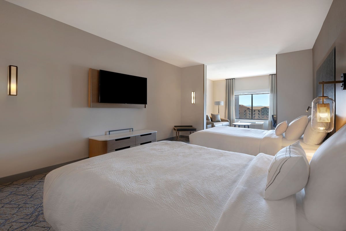 Habitación de hotel con dos camas, un televisor de pantalla plana montado en la pared, un escritorio con silla y una ventana que ofrece vistas a la ciudad.