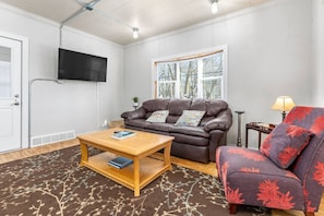 A living room with a brown leather sofa, a wooden coffee table, a red and purple armchair, a television mounted on the wall, and a window with a view of trees.