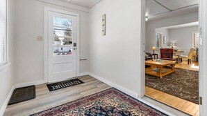 A hallway with a white door, a doormat, and a rug leading to a living room with a sofa, a coffee table, and a chair. - Stylish, Spacious Home: Walk Downtown, Fenced Yard on Washington St. (Appleton)