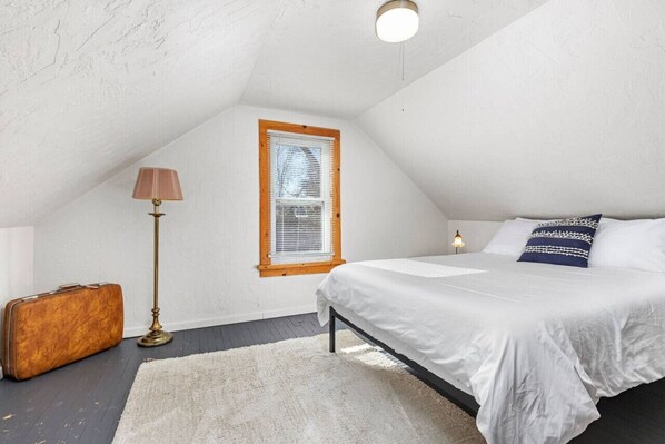 Une chambre à coucher avec un lit, une fenêtre avec des stores, une lampe de sol et une valise.