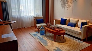 Een moderne hotelkamer met een houten vloer, een beige bank met blauwe kussens, een ronde salontafel, een klein bijzettafeltje en een grote waterdispenser.