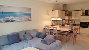A modern living room with a grey sofa, wooden dining table, and chairs. There is a kitchen area with cabinets and appliances.
