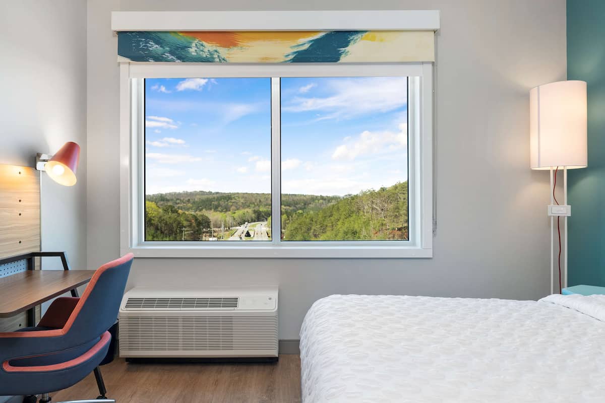 Une chambre d’hôtel avec une grande fenêtre, un bureau, une chaise, un climatiseur et un lit.