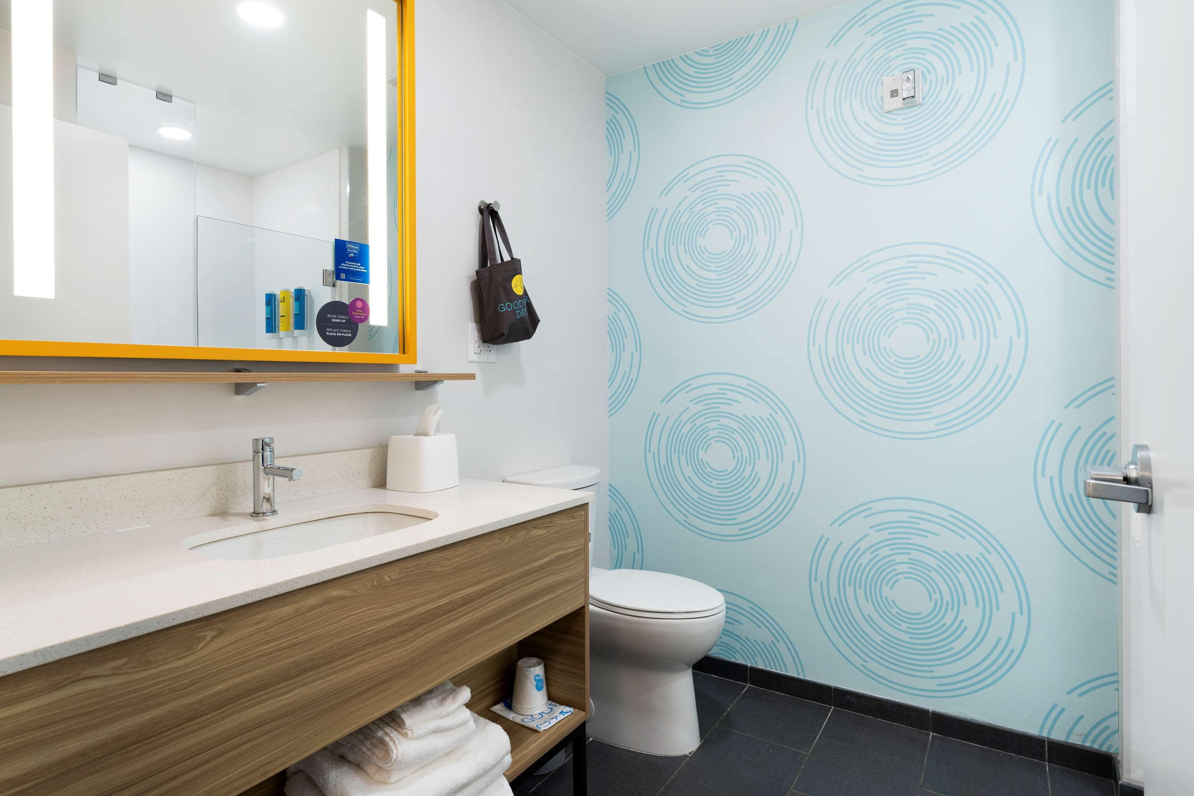A modern bathroom with a white sink, wooden vanity, and a toilet. There is a mirror with a yellow frame, and the walls have a light blue circular pattern.