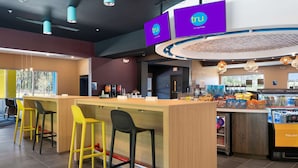 Un espace bar moderne avec un comptoir en bois, des tabourets de bar jaunes et noirs, et un présentoir réfrigéré contenant des collations.
