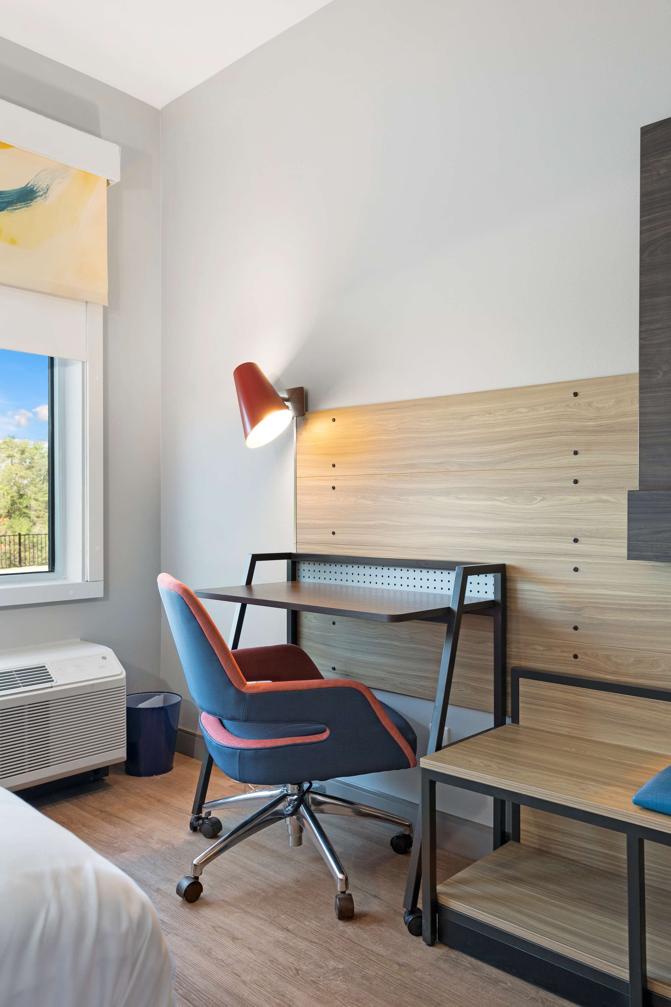 Habitación de hotel con cabecera de madera, un escritorio con silla, una lámpara y ventana con vistas a zonas verdes.