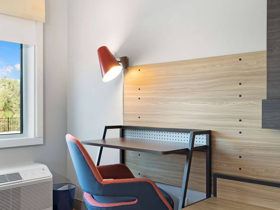 Habitación de hotel con cabecera de madera, un escritorio con silla, una lámpara y ventana con vistas a zonas verdes.