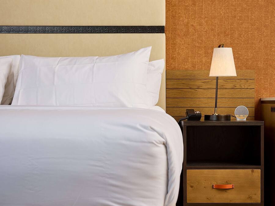 A neatly made bed with white linens, a wooden headboard, a bedside table with a lamp, and a nightstand with a phone and a small round mirror.