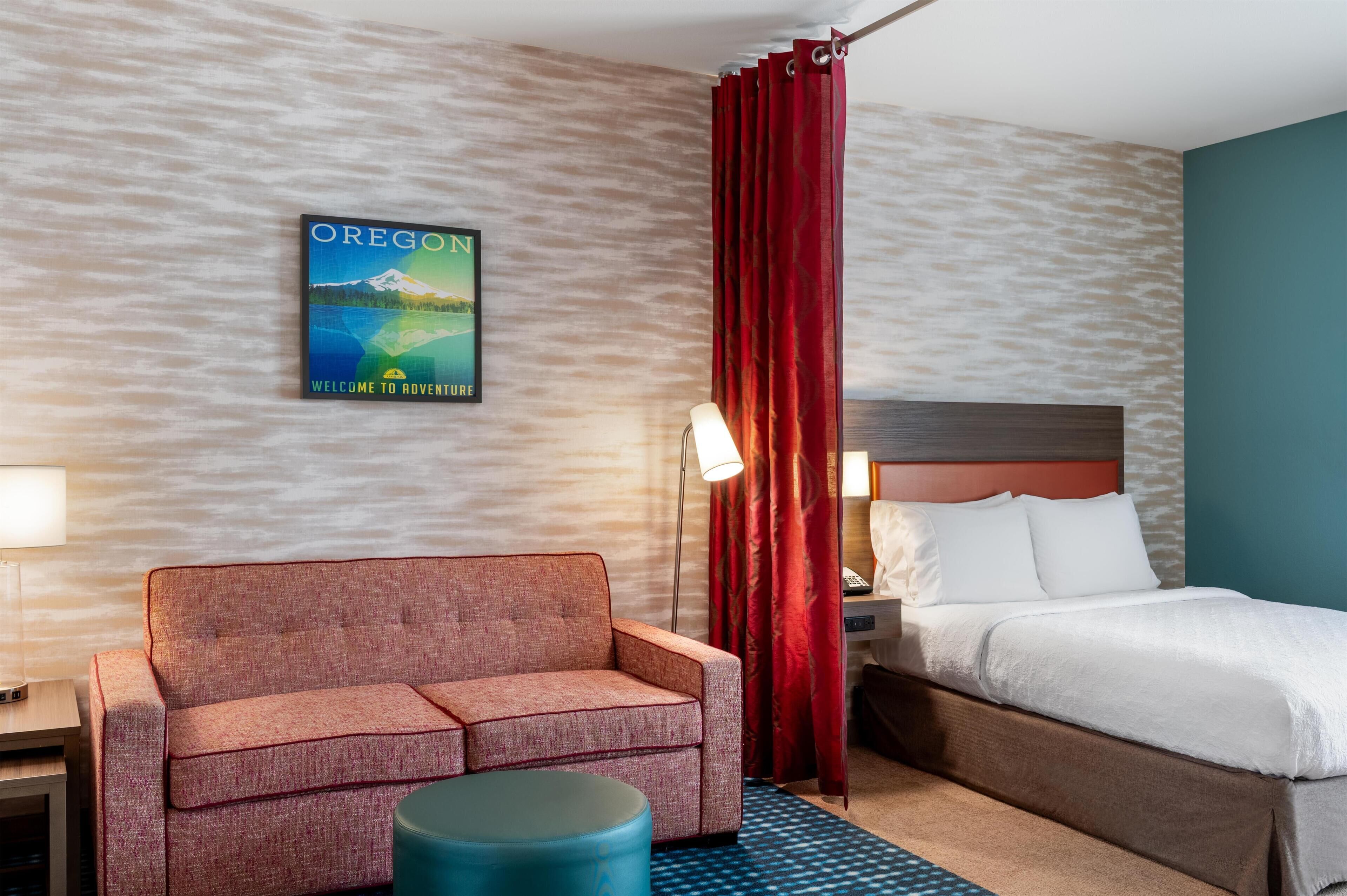 A hotel room with a bed, a sofa, a nightstand, a lamp, a red curtain, and a wall poster.