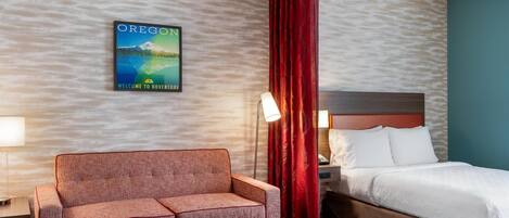 A hotel room with a bed, a sofa, a nightstand, a lamp, a red curtain, and a wall poster.
