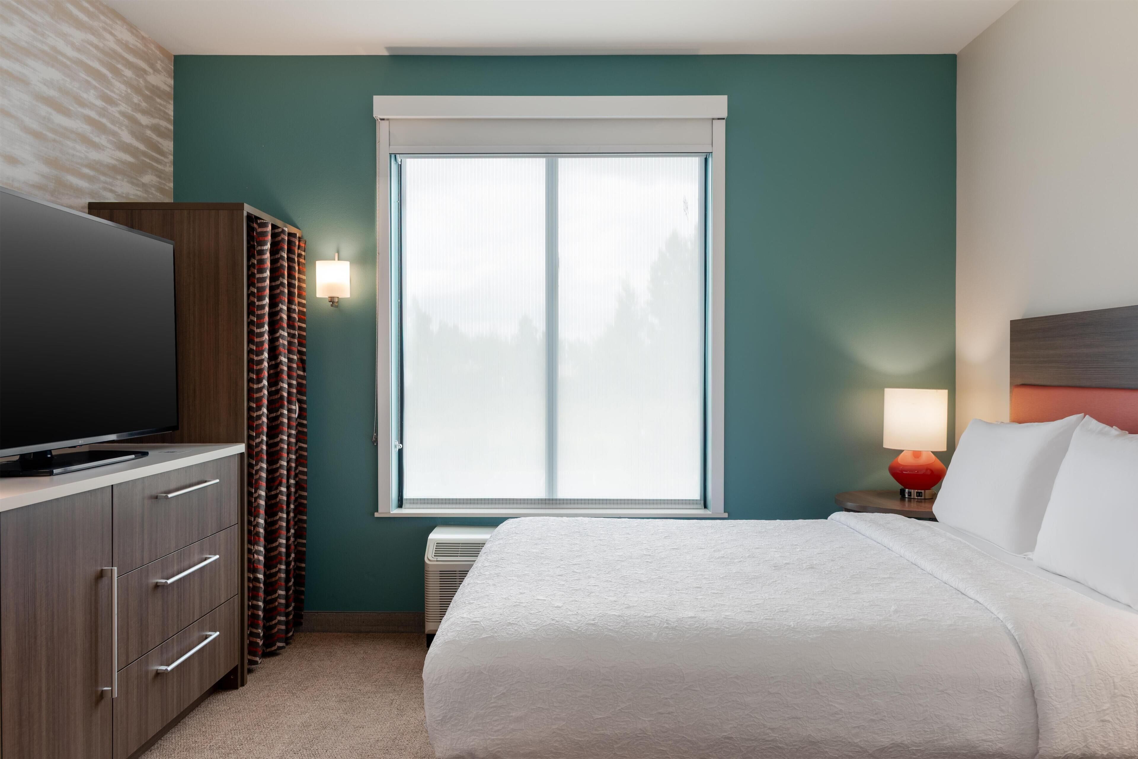 Habitación de hotel con una cama grande, un televisor sobre un mueble de madera, una ventana con cortinas y una mesita de noche con lámpara.