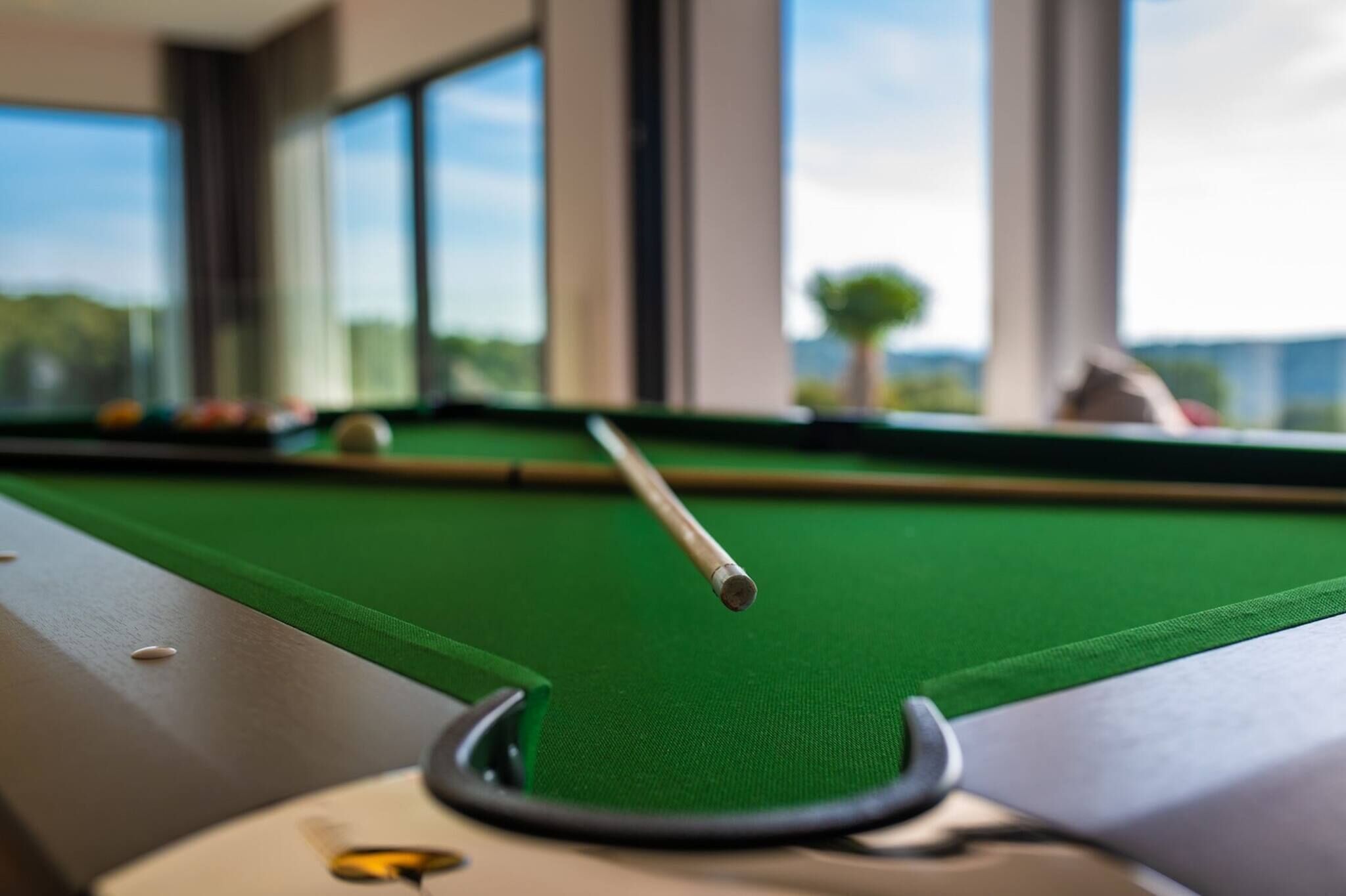 A pool table with a cue stick in motion, a green felt surface, and a window with a view of trees and buildings.