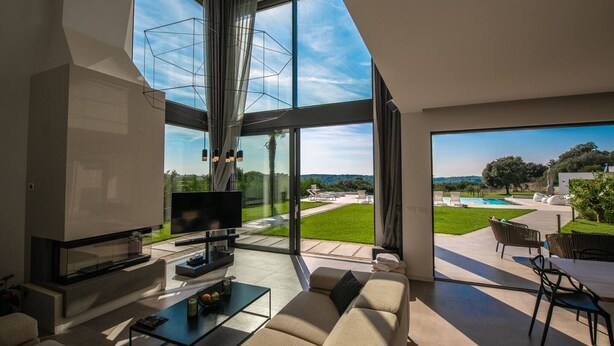 A modern living room with a large TV, comfortable seating, and a view of a pool and garden.