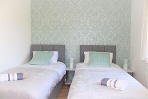 Two single beds with grey headboards and white mattresses, set against a light green damask backdrop.