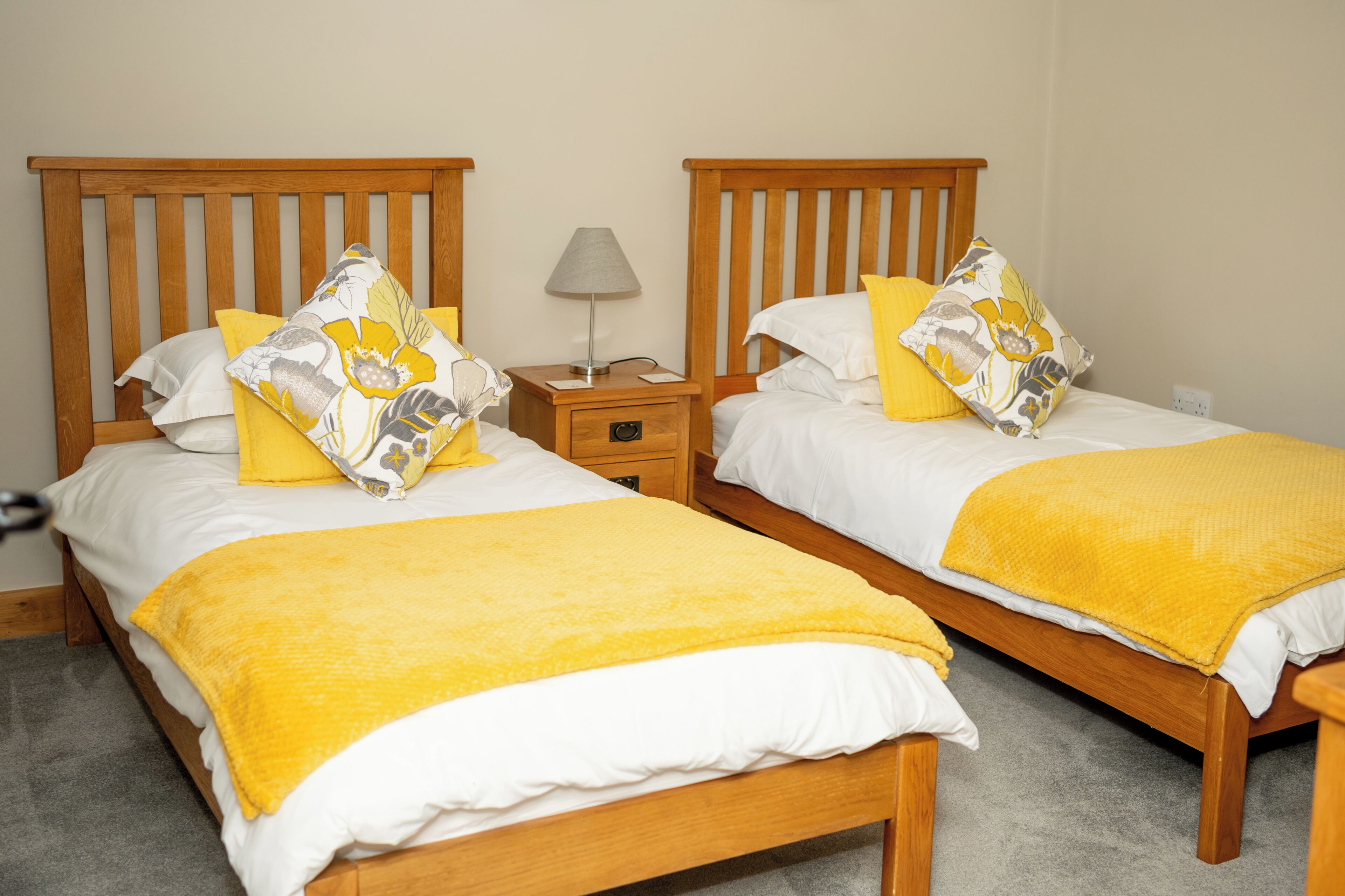 Two wooden beds with white bedding and yellow accents, a bedside table with a lamp, and a grey carpeted floor.