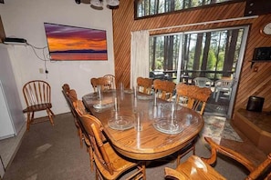 A dining area with a large wooden table and chairs, a painting of a sunset, and a view of trees through large windows. - Great wooded location within Walking Distance of Heavenly! 1360K (South Lake Tahoe)