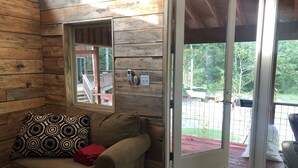 Interior de una cabaña de madera con sofá, cojín estampado y puerta corredera de cristal que da a una terraza exterior.