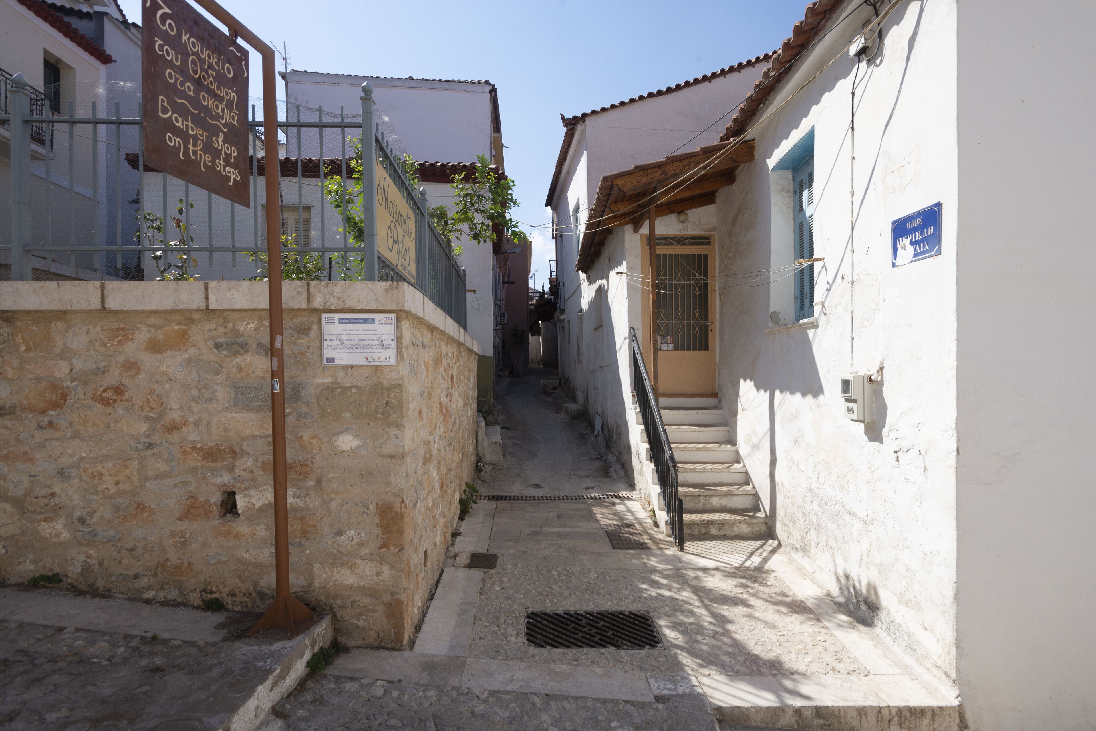 Un estrecho callejón con paredes de piedra, una reja metálica y una escalera que conduce a un edificio con un letrero azul.