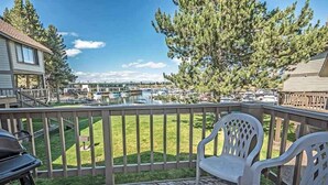 A wooden deck with two white plastic chairs, a grill, and a view of a marina with boats and a building in the background.