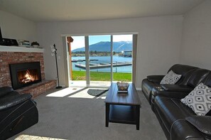 A living room with a fireplace, a black leather sofa, a coffee table, and a view of a lake and mountains through large glass doors.