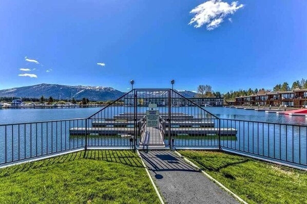 A lakeside view with a bridge leading to a dock, surrounded by mountains and a clear sky.