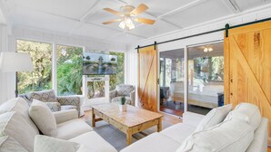 A spacious living room with a large sofa, a coffee table, and a TV mounted on the wall. There is a sliding barn door leading to an outdoor area.