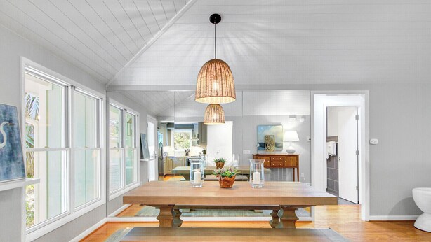 A spacious dining area with a large wooden table, a hanging woven light fixture, and a view into the kitchen through large windows.