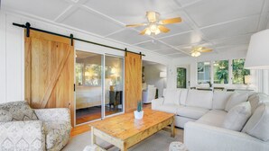 A spacious living room with a large sliding glass door leading to a bedroom, a white sofa, a wooden coffee table, and a ceiling fan.