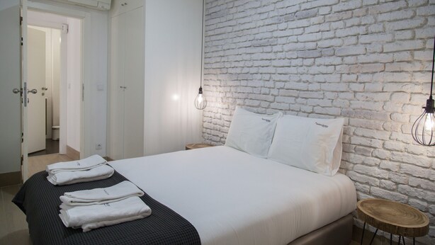 A hotel room with a brick wall, a neatly made bed with white linens, and a wooden side table. - FLH Alfama Traditional Flat (Lisbon)