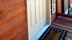 Entrada de una cabaña de madera con una puerta blanca, un cartel de bienvenida y un felpudo.