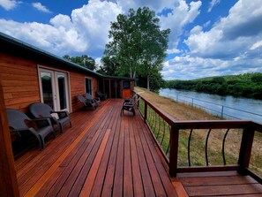 Eine Holzterrasse mit Außenmöbeln, Blick auf einen Fluss und ein überdachter Bereich mit einer Tür.