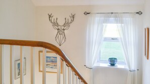 A staircase with a wooden handrail, a wall-mounted deer head decoration, a window with sheer curtains, and a framed picture on the wall. - 4 bedroom accommodation in Nairn (Nairn)