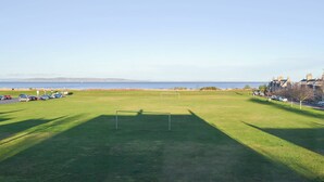 A grassy field with a goalpost, a parking area with cars, and a view of the sea and buildings in the background. - 4 bedroom accommodation in Nairn (Nairn)