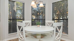A round dining table with white chairs, centered between two windows with blinds. - Golf Cart ~ Screened Patio ~ Golf Course Views (Miramar Beach)