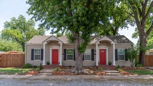 A two-story house with a central tree, a red door, and a small front yard with a wooden fence. - The Cozy Chickasha | Fiber | Extra Parking | W/D (Chickasha)